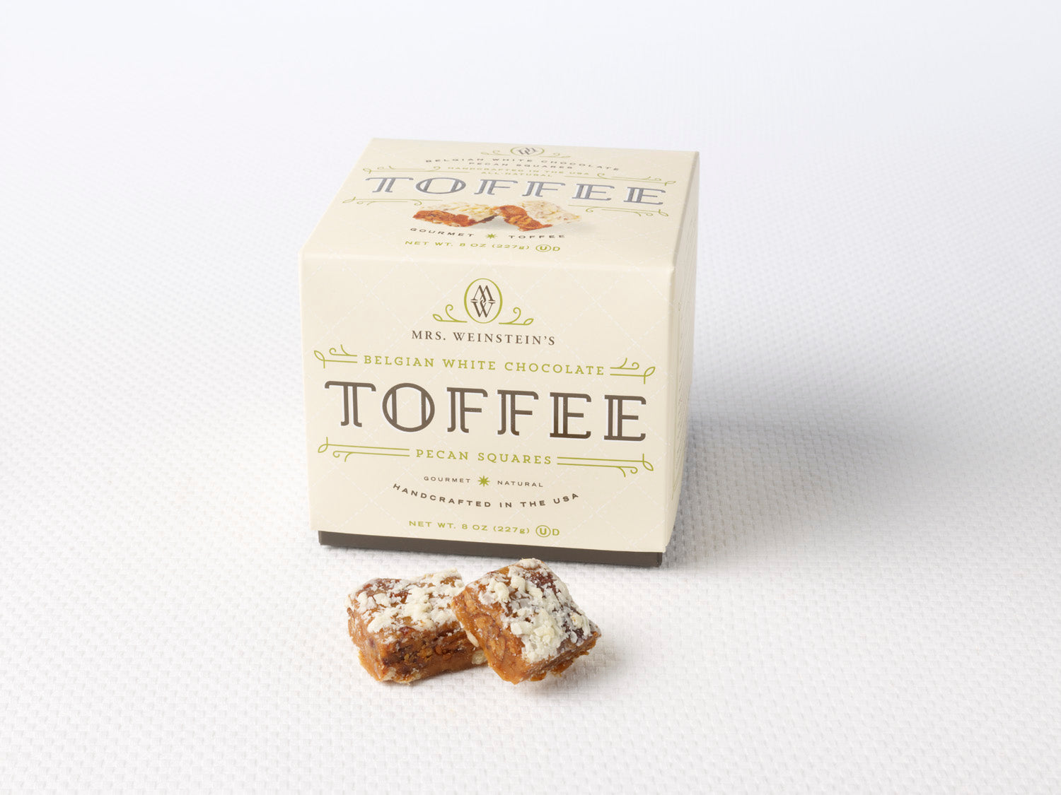 A box of Belgian white chocolate pecan toffee squares with two pieces of toffee in front of the box.